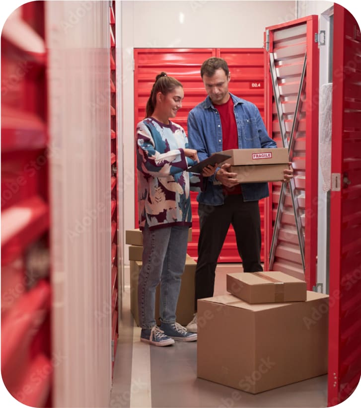 A girl and a boy holding a box in front of an open red door of a self storage.